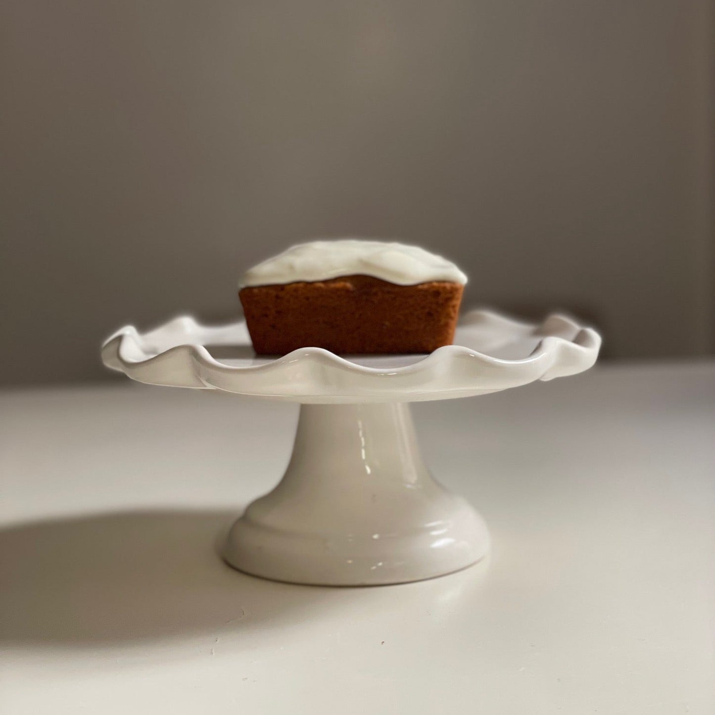 Small cake with white frosting on a white ceramic stand against a plain background